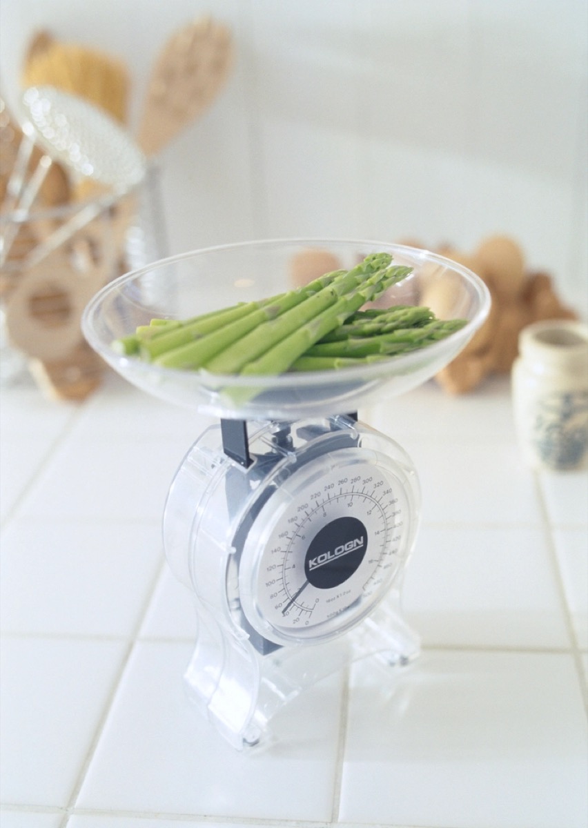 A glass bowl of fresh green asparagus spears sitting on a kitchen scale on a white countertop, with wooden utensils blurred in the background.