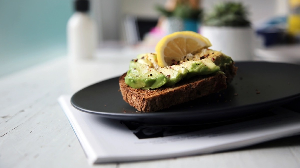 A slice of avocado toast on wholegrain bread with chilli flakes and a lemon wheel on a black plate