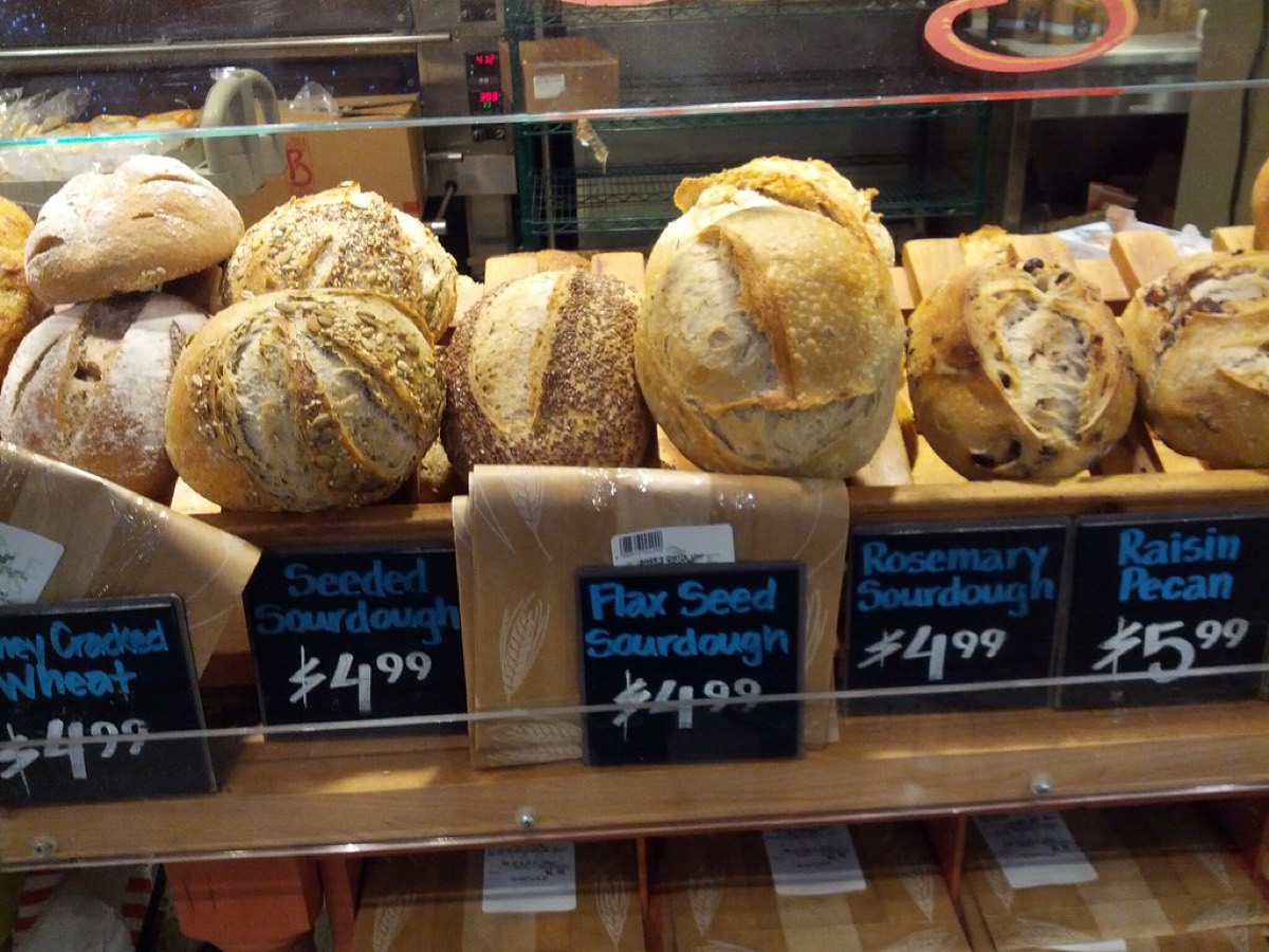 Rows of fresh sourdough and specialty bread loaves on a wooden bakery shelf with handwritten price signs