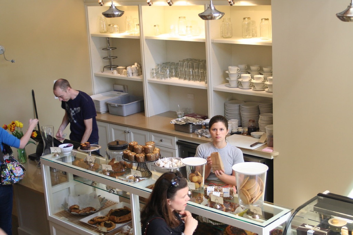 A bakery staff member holds a wrapped loaf behind a glass pastry case filled with croissants and tarts while a customer browses the display at the counter.