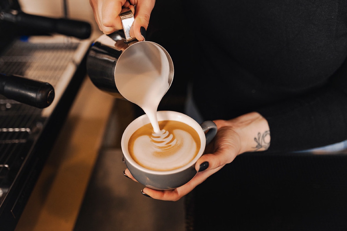 A barista's hands pouring steamed milk from a jug into a cup to form latte art beside an espresso machine