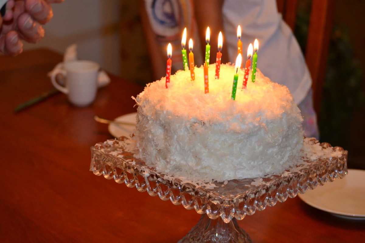 A frosted coconut cake with several lit birthday candles on a wooden table