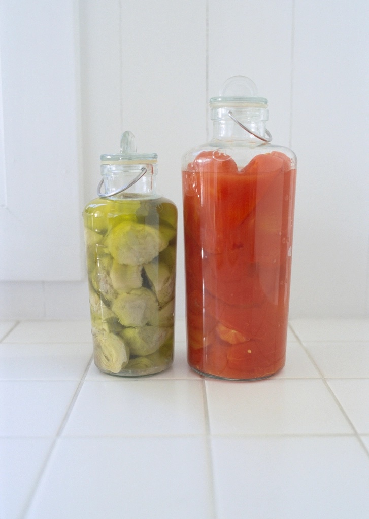 Two clear glass flip-top jars on a white tiled surface, one filled with pickled vegetables and one filled with red tomato sauce
