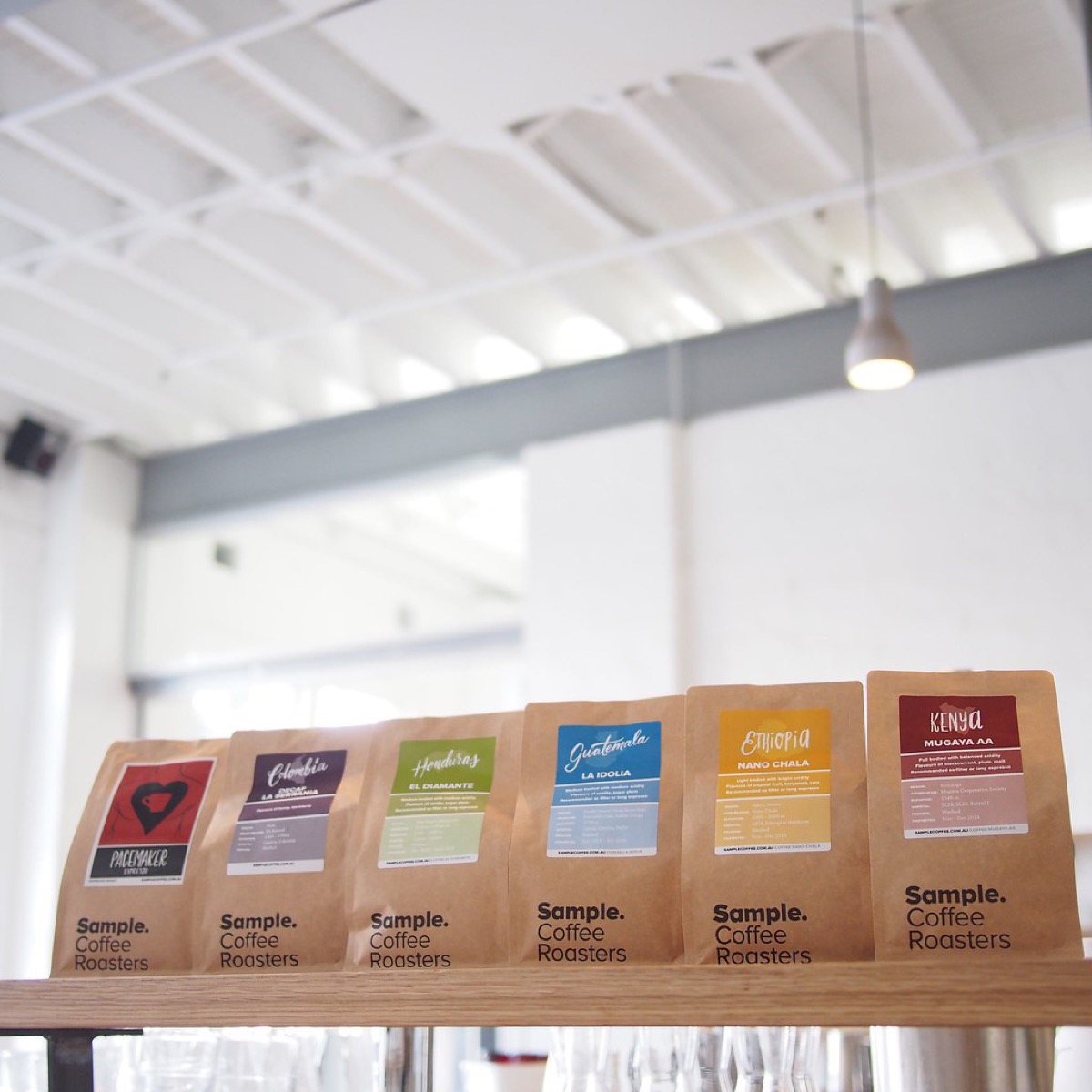 A row of kraft paper coffee bags on a wooden shelf, each with a different coloured label and the same wordmark logo, showing a consistent brand identity across a product range.