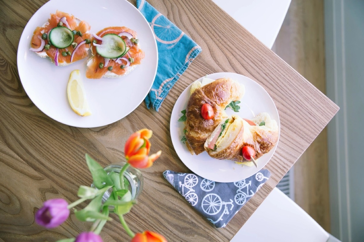 Overhead view of a smoked salmon bagel and a stuffed croissant sandwich on white plates on a wood table with tulips