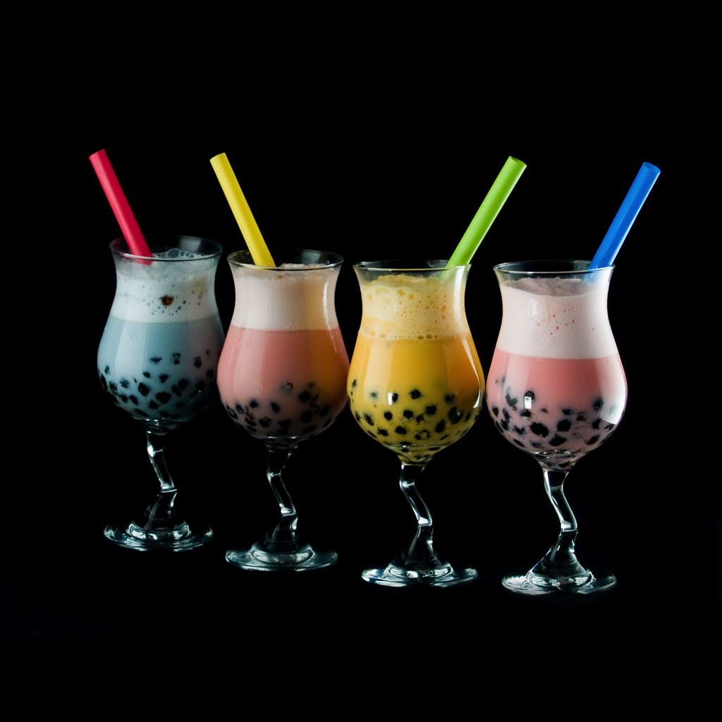Four bubble tea drinks in stemmed glasses with colourful straws and visible tapioca pearls, set against a black background