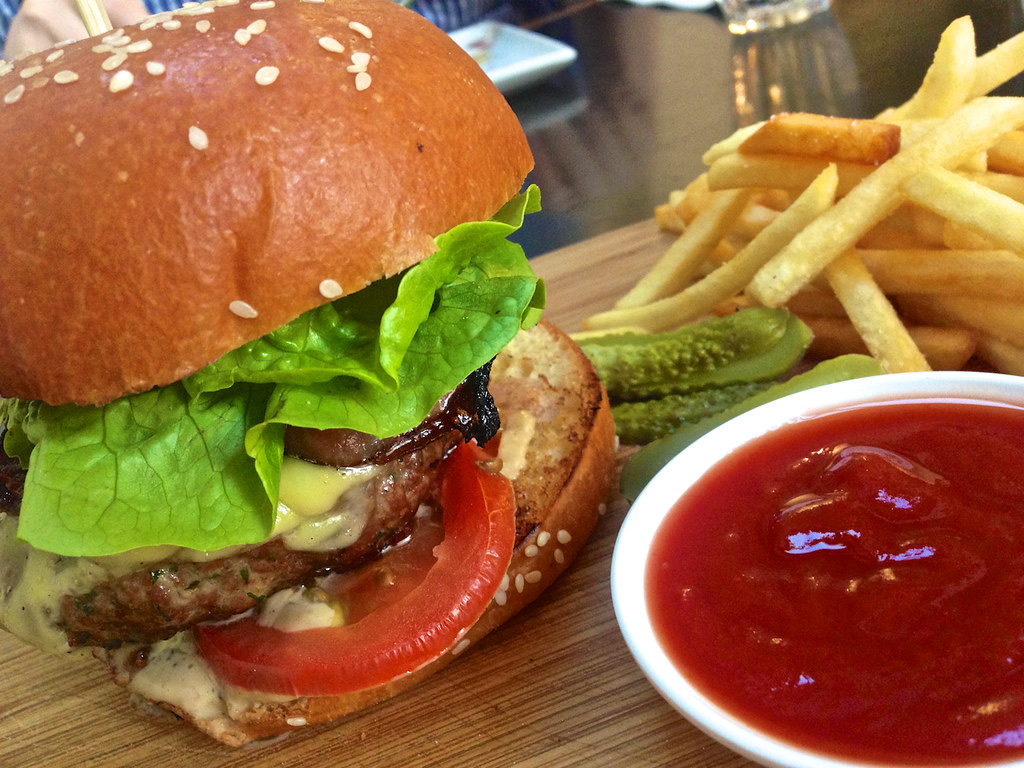Close-up of a stacked burger with melted cheese, lettuce and tomato beside a pile of golden fries and a bowl of ketchup