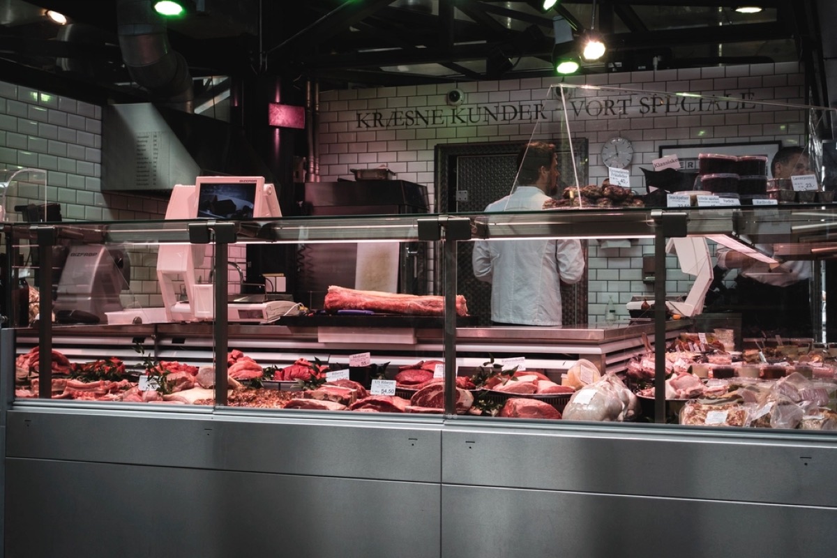 A long refrigerated butcher display case full of fresh cuts of meat, with staff working behind it in a tiled market hall butchery