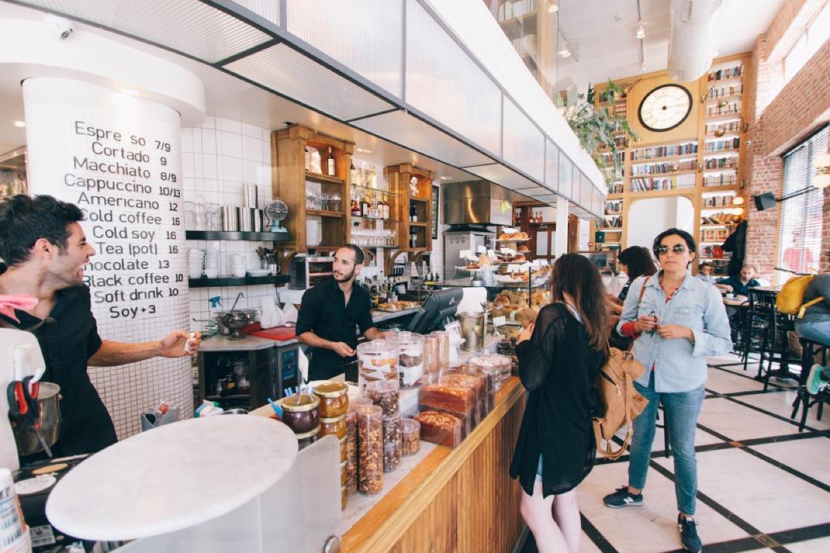 A barista stands at a cafe counter beside a point of sale terminal and pastry display case while two customers wait to order.