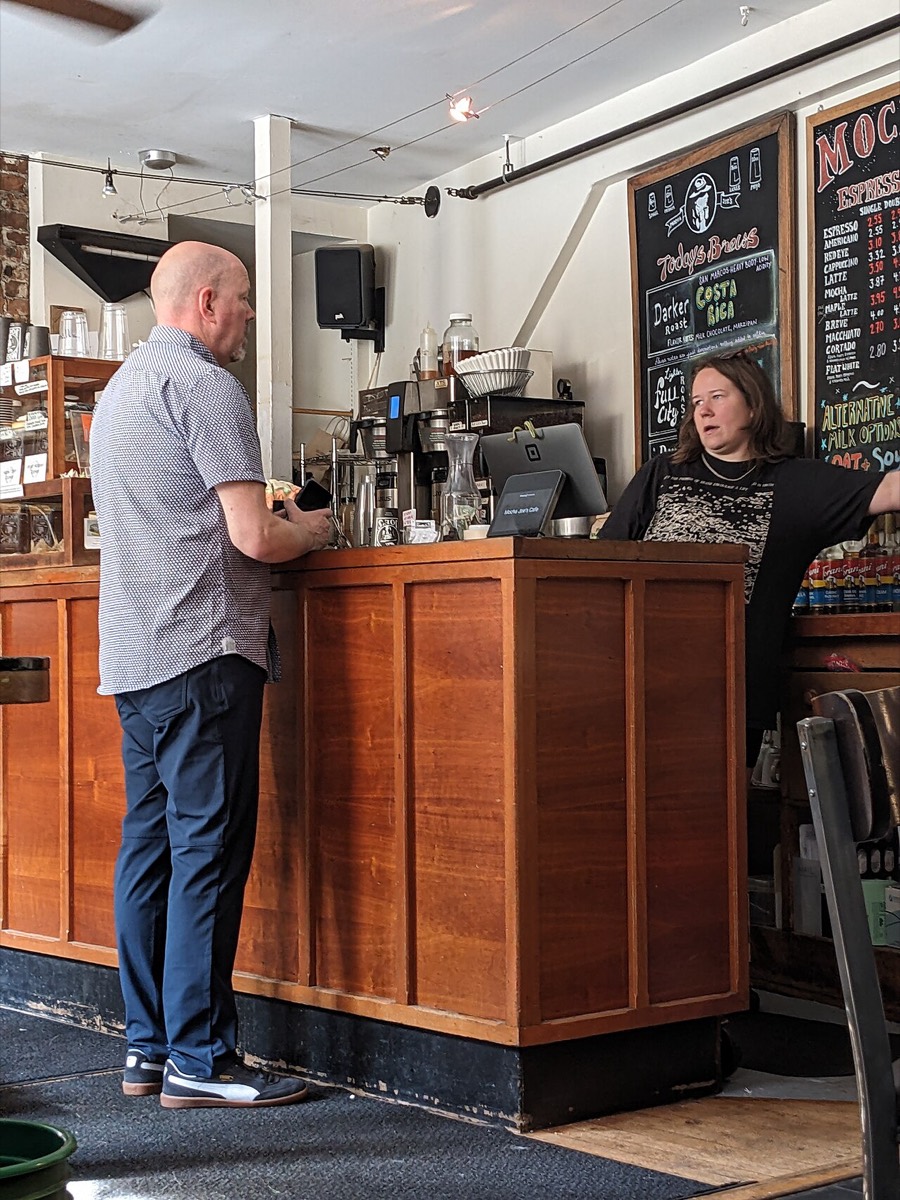 A customer talking with a barista across a wooden counter inside a sunlit cafe with a chalkboard menu
