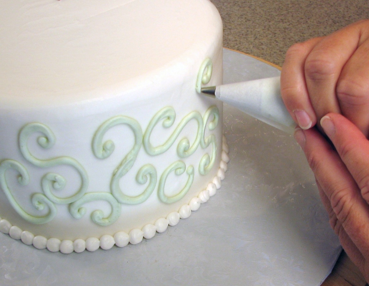 A baker's hand pipes pale green buttercream scrollwork onto the fondant side of a white cake with a beaded border.