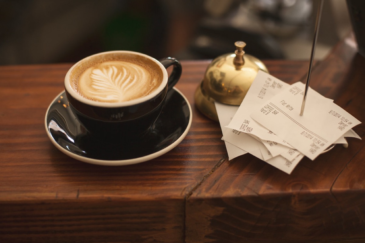 A single black cappuccino cup with leaf-pattern latte art sitting on its saucer on a wooden cafe counter, next to a stack of order receipts on a spike.