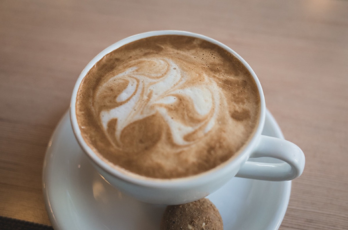 A cappuccino with swirled latte art in a white cup and saucer on a light wooden table