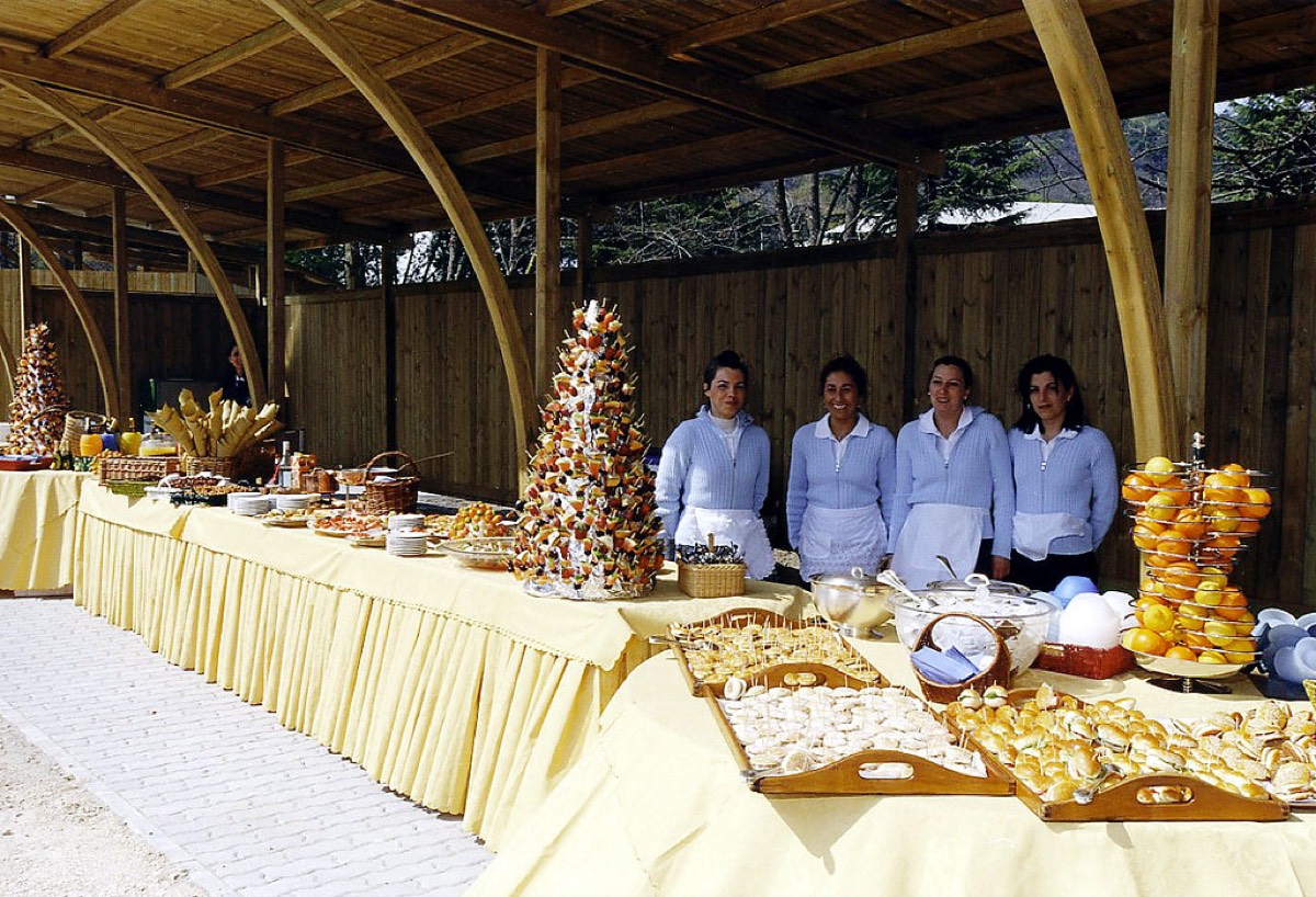 A long buffet table laid with catering trays of canapes and appetizers under a wooden pavilion, with catering staff in white uniforms standing behind it.