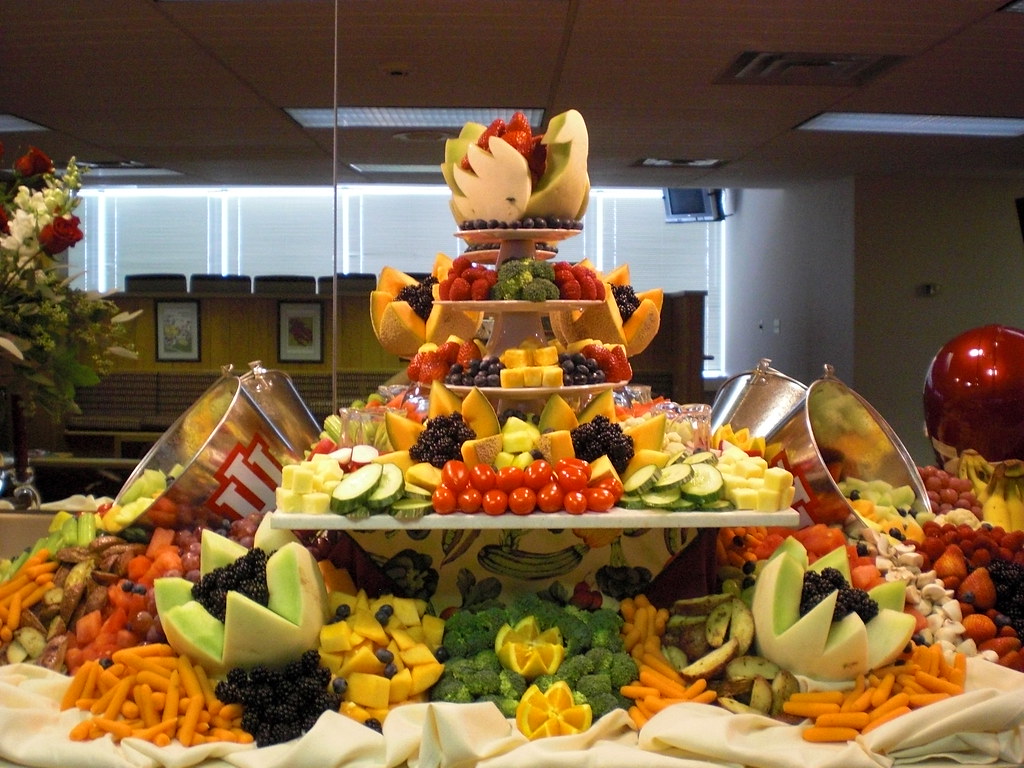 A tiered catering display piled with fresh fruit and vegetables — melon, berries, carrots, broccoli and citrus — at an indoor event