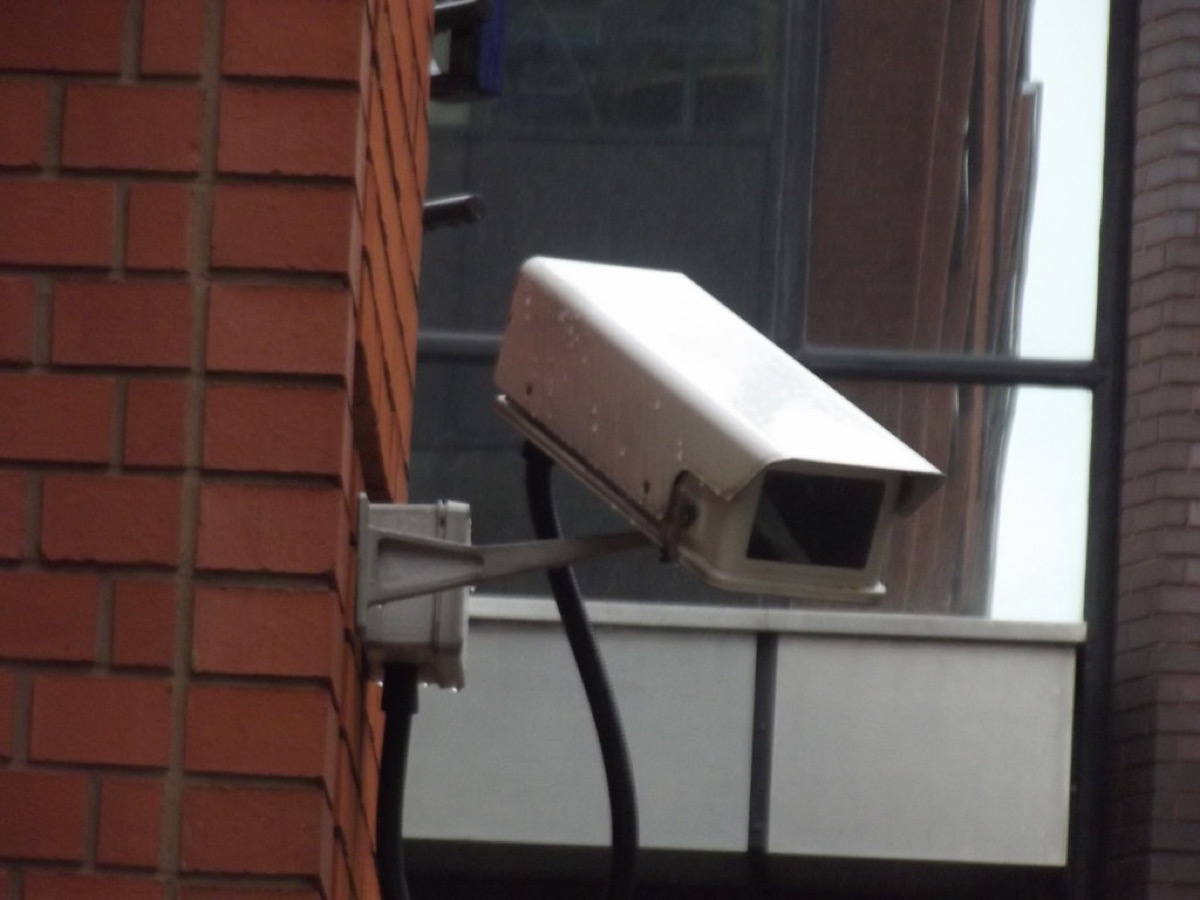 A white CCTV security camera mounted on a bracket against a red brick wall, pointed toward a storefront entrance.