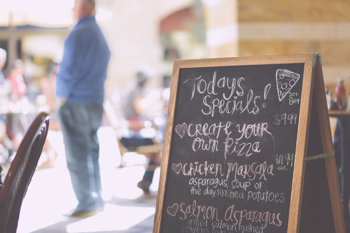 A handwritten chalkboard menu reading Todays Specials, listing Create Your Own Pizza, Chicken Marsala with asparagus soup of the day and mashed potatoes, and Salmon Asparagus, with a diner blurred in the background.