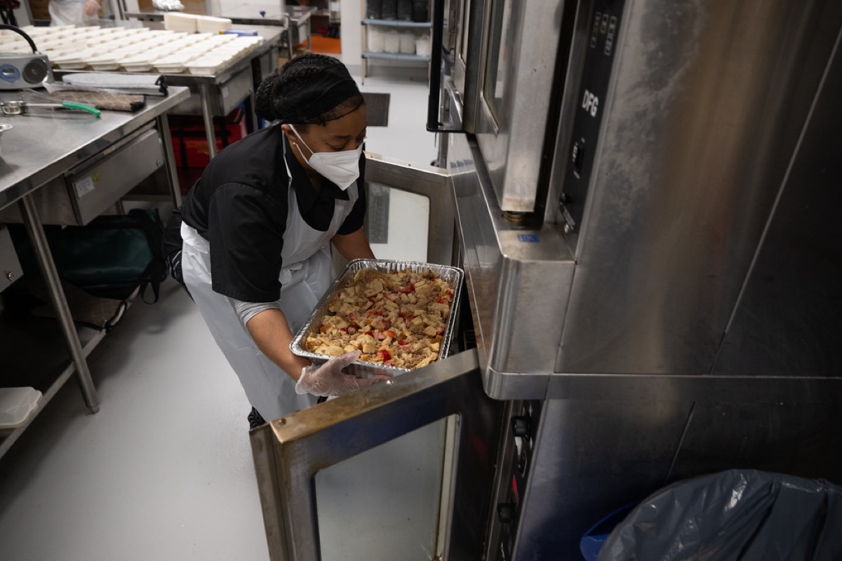 A kitchen worker in a face mask and gloves lifts a full foil catering tray of baked food out of a commercial oven.