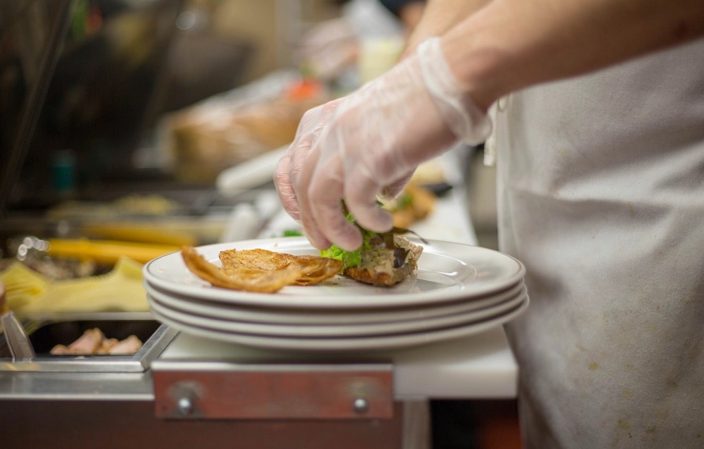 A chef wearing a clear disposable glove places food onto a stack of white plates in a professional kitchen.