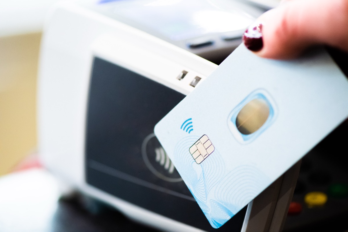A close-up of a hand holding a chip and contactless payment card in front of a blurred card reader terminal.