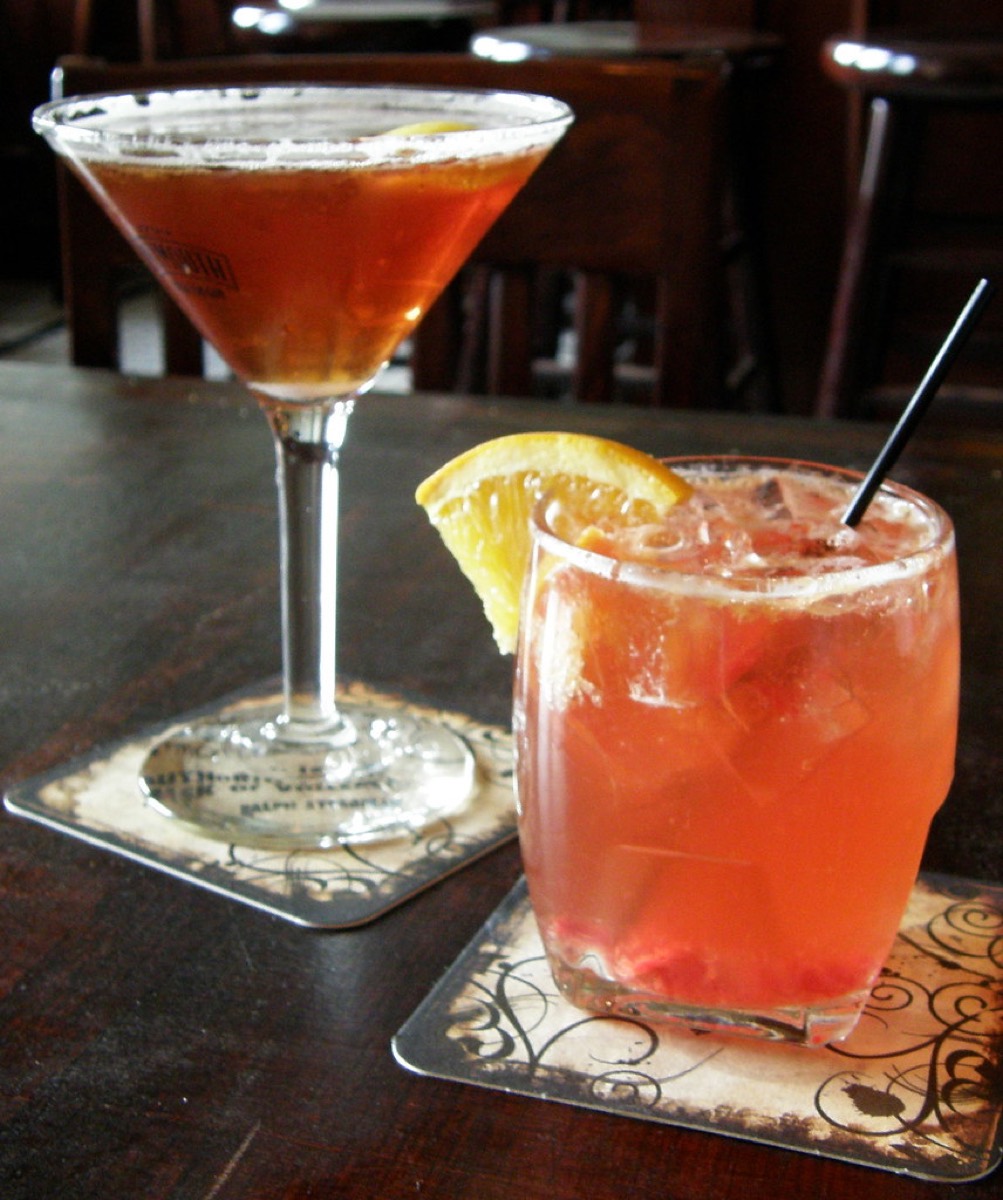 A martini and an orange cocktail with an orange slice garnish and straw, served on coasters at a dark wood bar table.