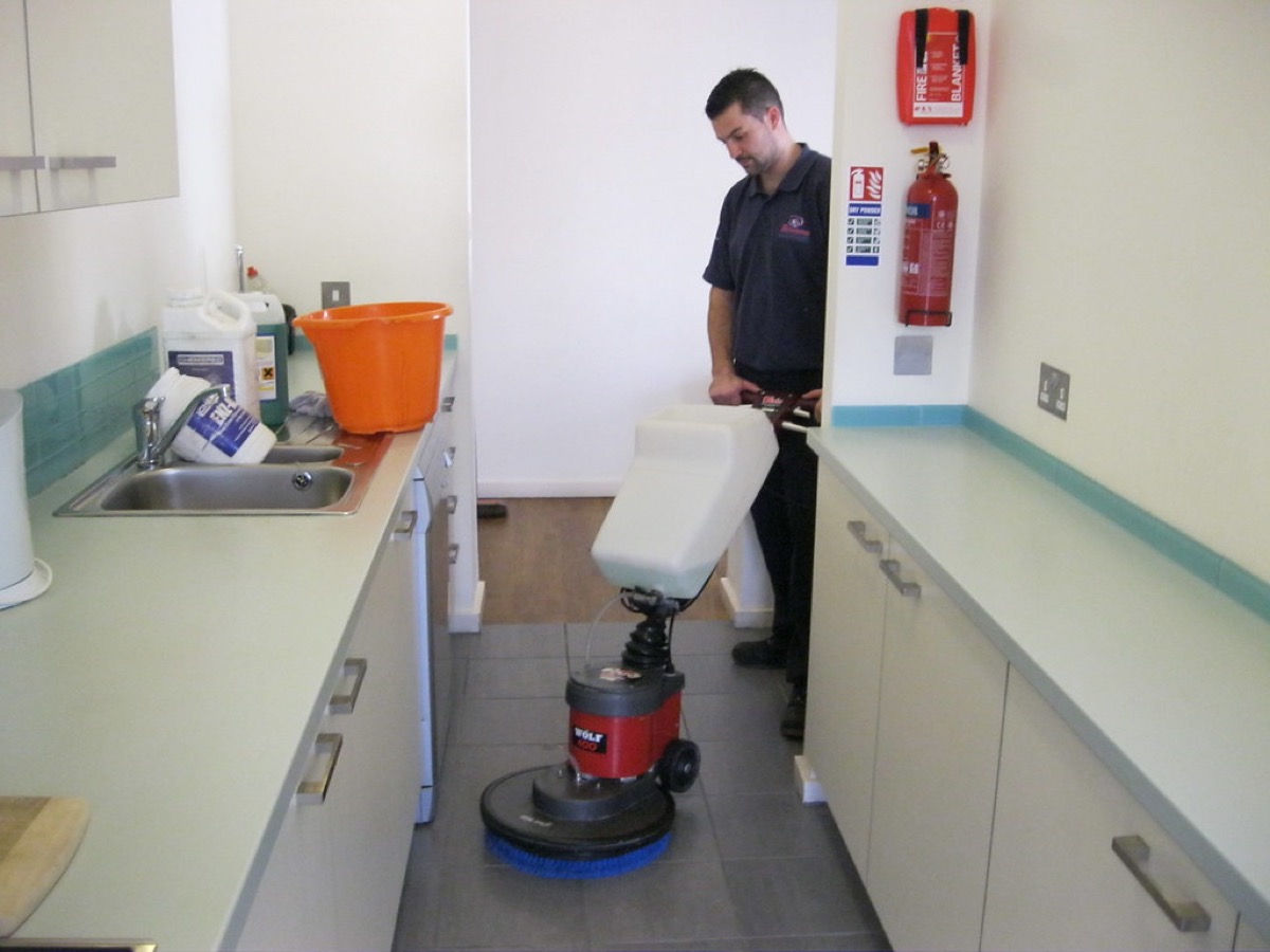A staff member in a commercial kitchen runs a large floor buffing machine across the tiles, with cleaning supplies on the counter and a fire safety station on the wall behind him.
