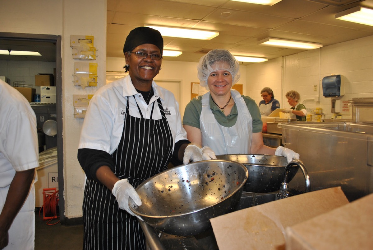 Two commercial kitchen staff wearing hairnets, gloves and aprons smile while washing produce in stainless steel bowls, with more staff working in the background.