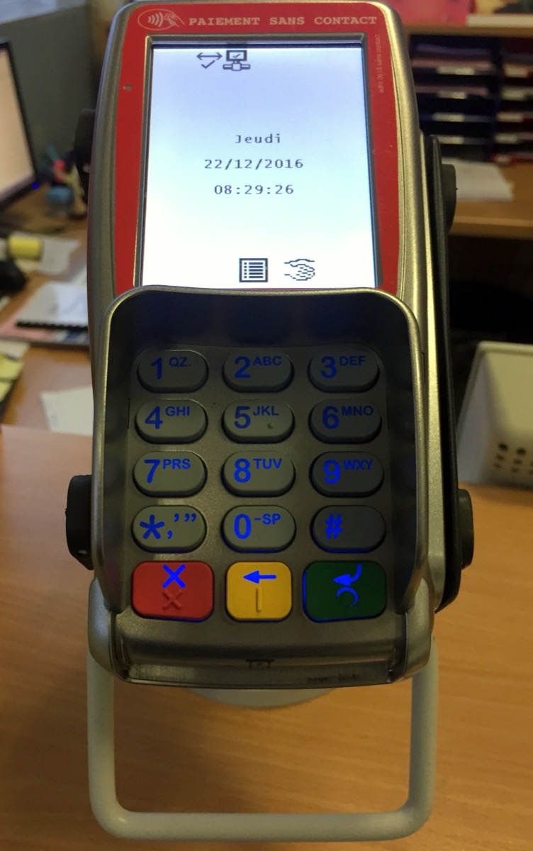 A handheld contactless payment terminal with a numeric keypad and a screen showing the date and time, standing on its charging cradle on a wooden desk.