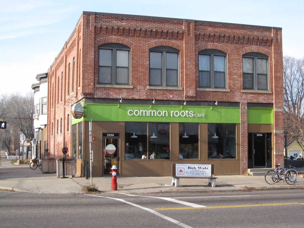 A brick corner building with a green awning reading Common Roots Cafe, large storefront windows, a bench, a fire hydrant, and parked bicycles on the sidewalk.