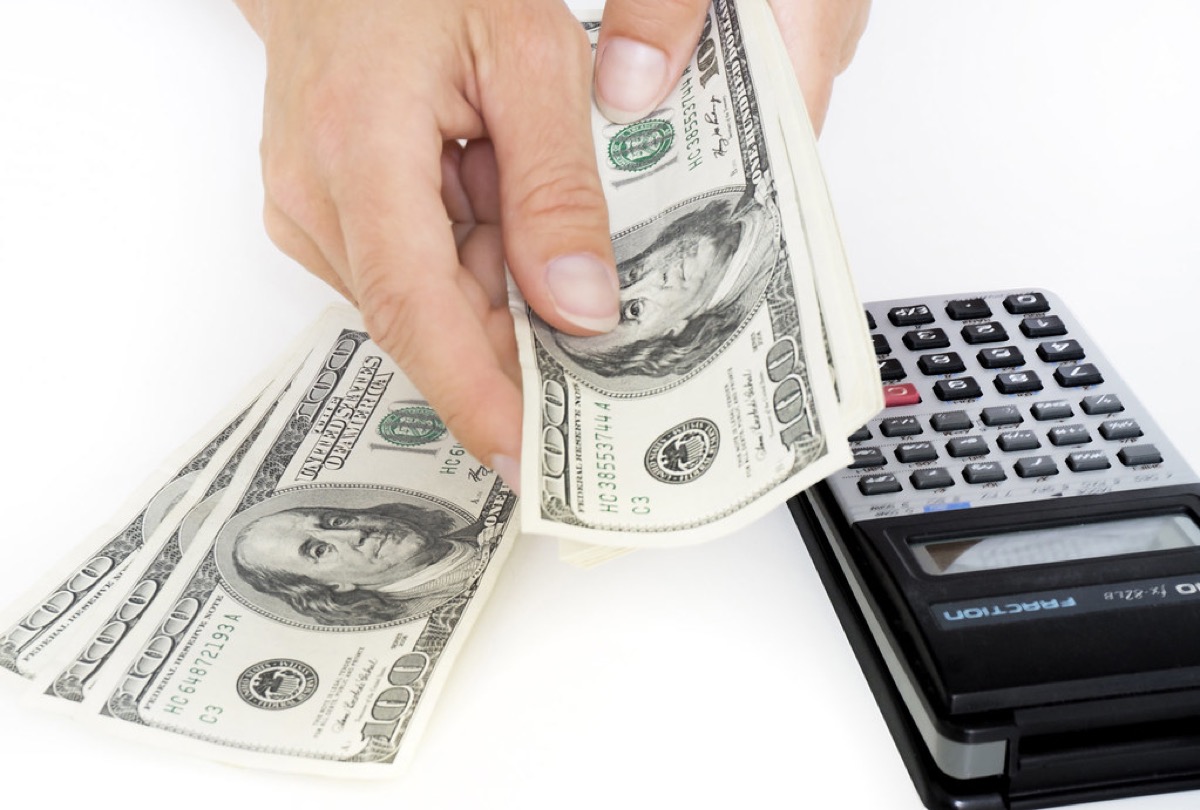 A pair of hands fans out several hundred dollar bills next to a calculator on a white surface.