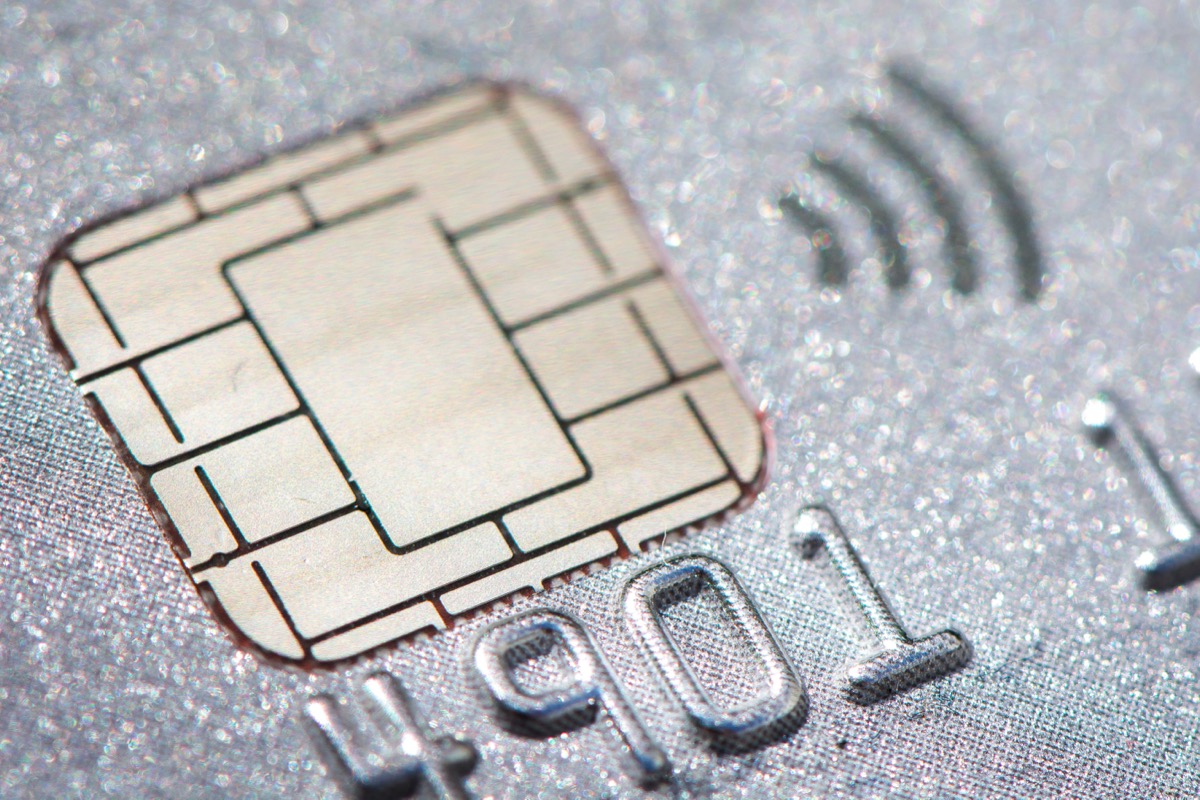 Extreme macro photo of a credit card's gold EMV chip and the four contactless payment arcs embossed on a silvery card, with a few stray digits visible but no full card number.