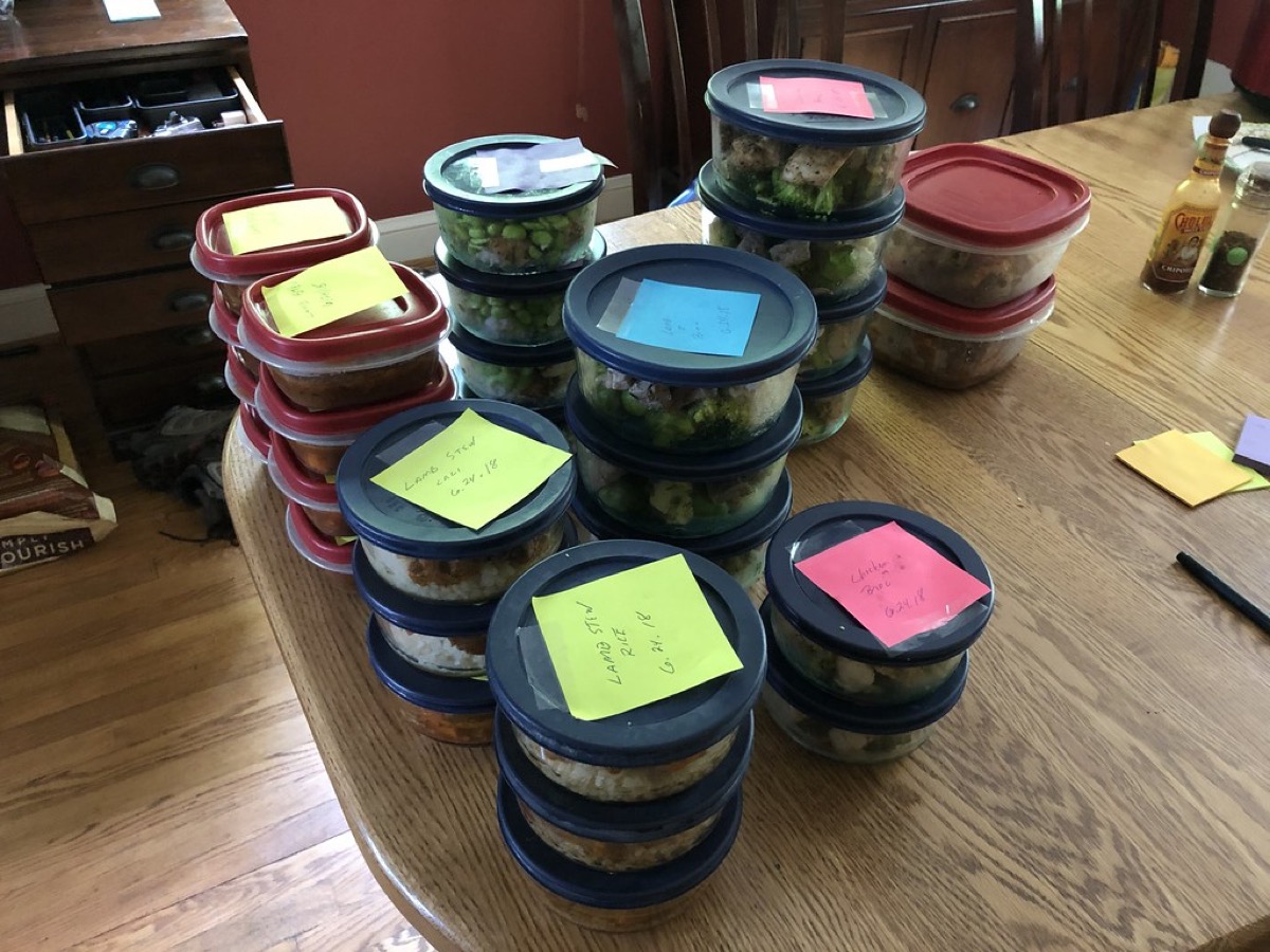 Stacked plastic food storage containers on a table, each topped with a handwritten sticky note label naming the contents and the date it was prepared.