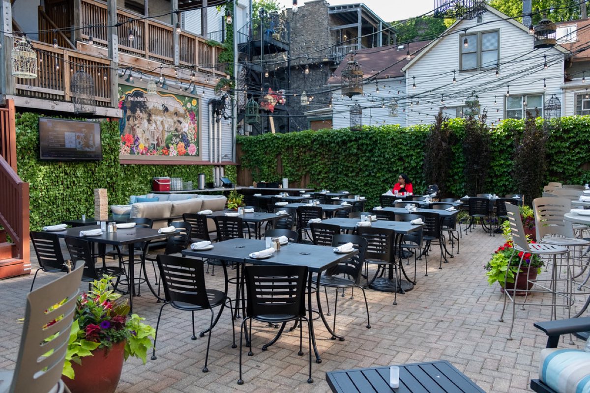 A sunlit outdoor restaurant patio with rows of tables set with white napkins and salt and pepper shakers, string lights and an ivy covered wall, with one guest seated at a table.
