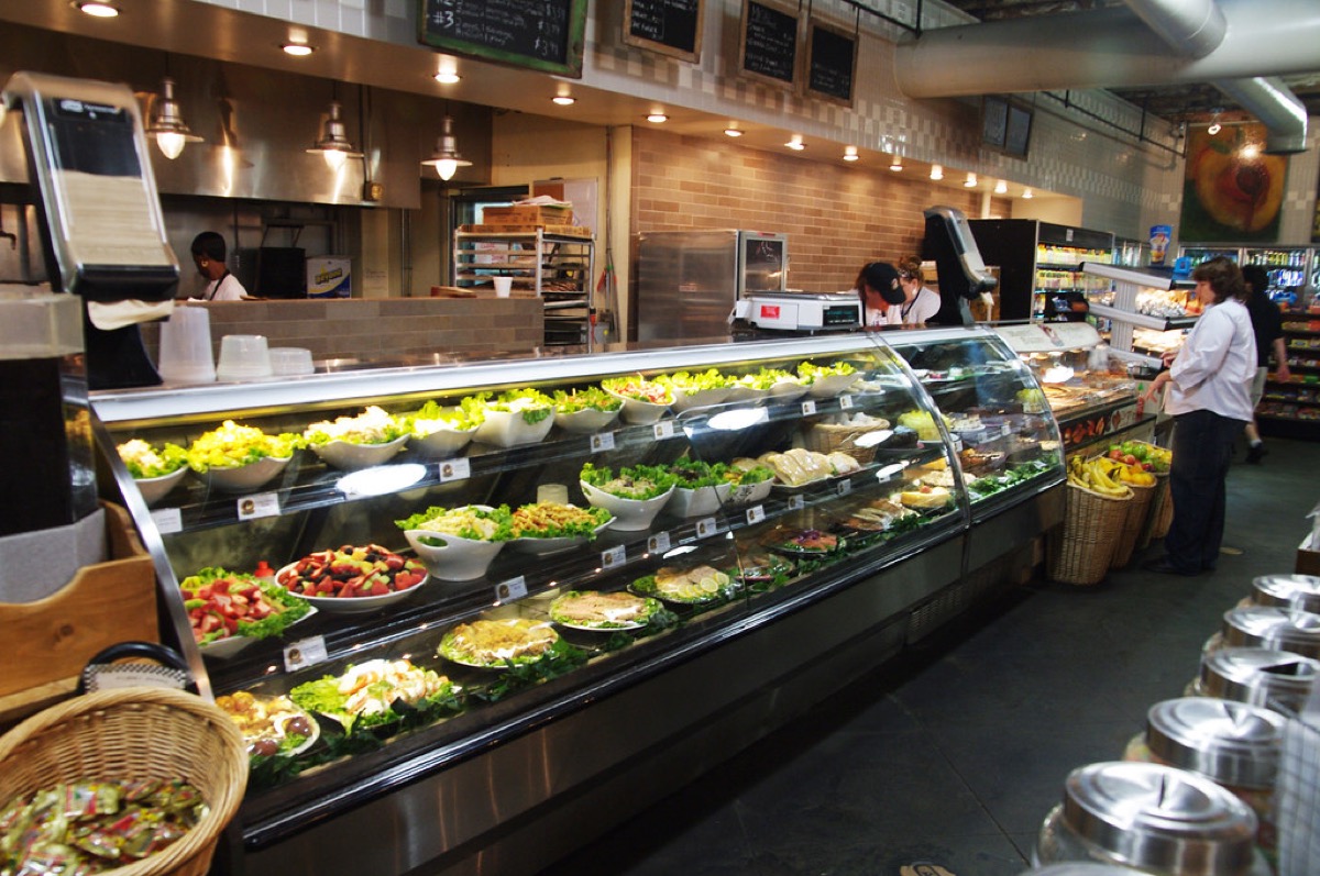 A refrigerated deli display case filled with prepared salads and cold dishes, with staff working behind the counter
