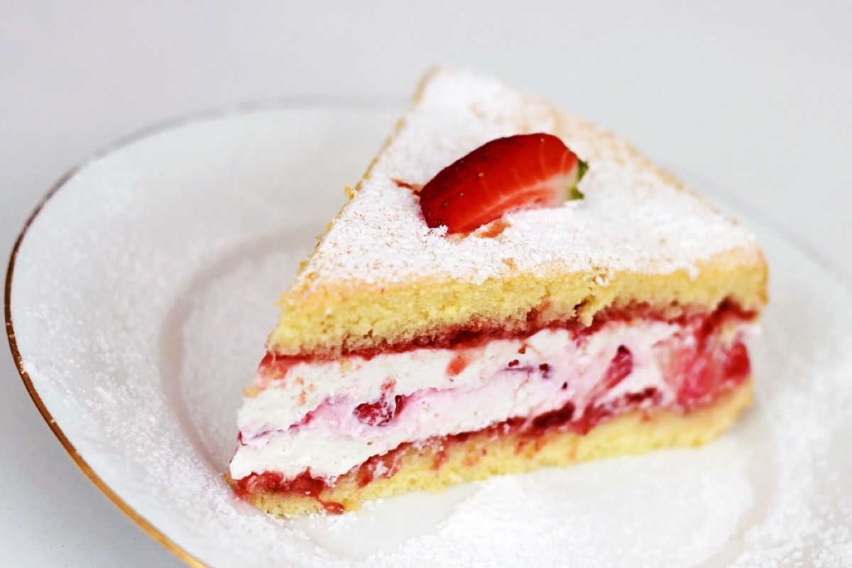A slice of strawberry sponge cake with cream filling, dusted with powdered sugar and topped with a fresh strawberry, on a white plate with gold rim