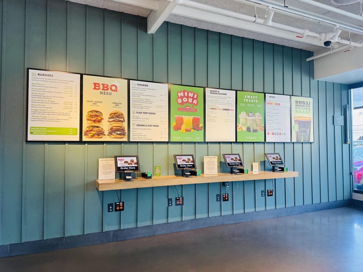 A wall of bright digital menu board screens showing burgers, chicken and drinks above a row of ordering kiosks in a quick-service restaurant