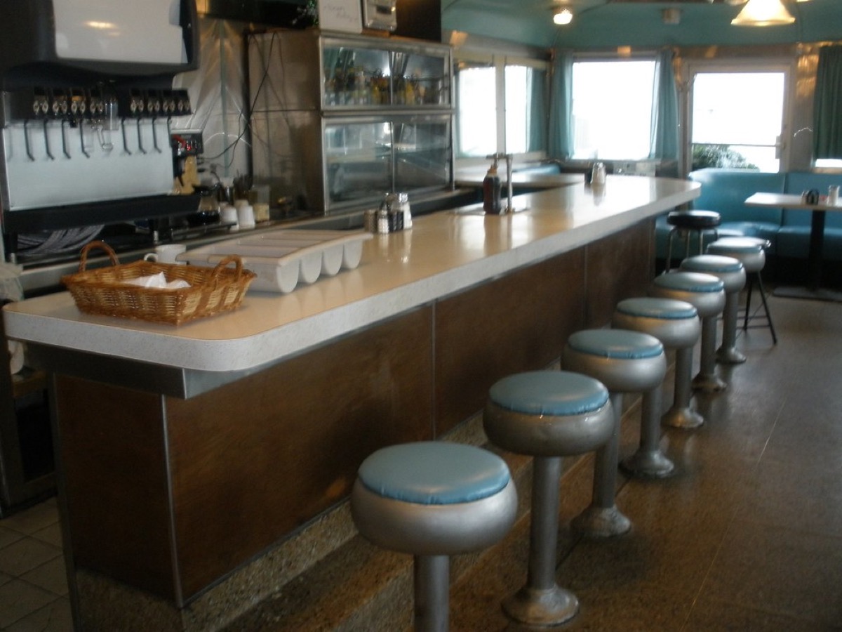 A classic diner counter with a row of blue vinyl stools, coffee machine and booth seating behind