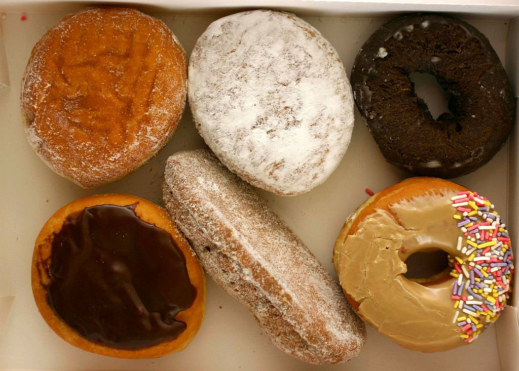 An overhead view of six assorted donuts in a box, including powdered, chocolate glazed, and sprinkle-topped varieties