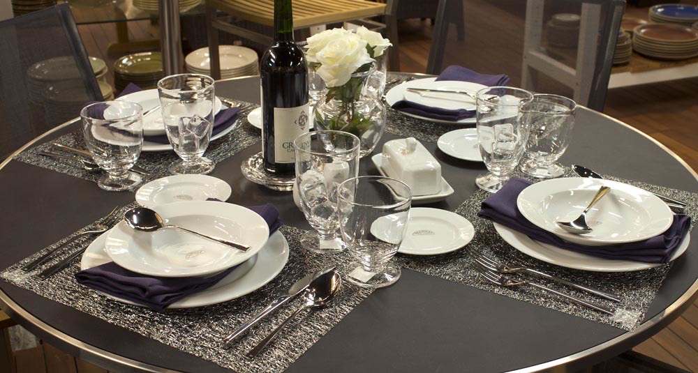Two formal place settings on a round table with white plates, folded purple napkins, wine glasses, and a wine bottle beside a white rose centerpiece