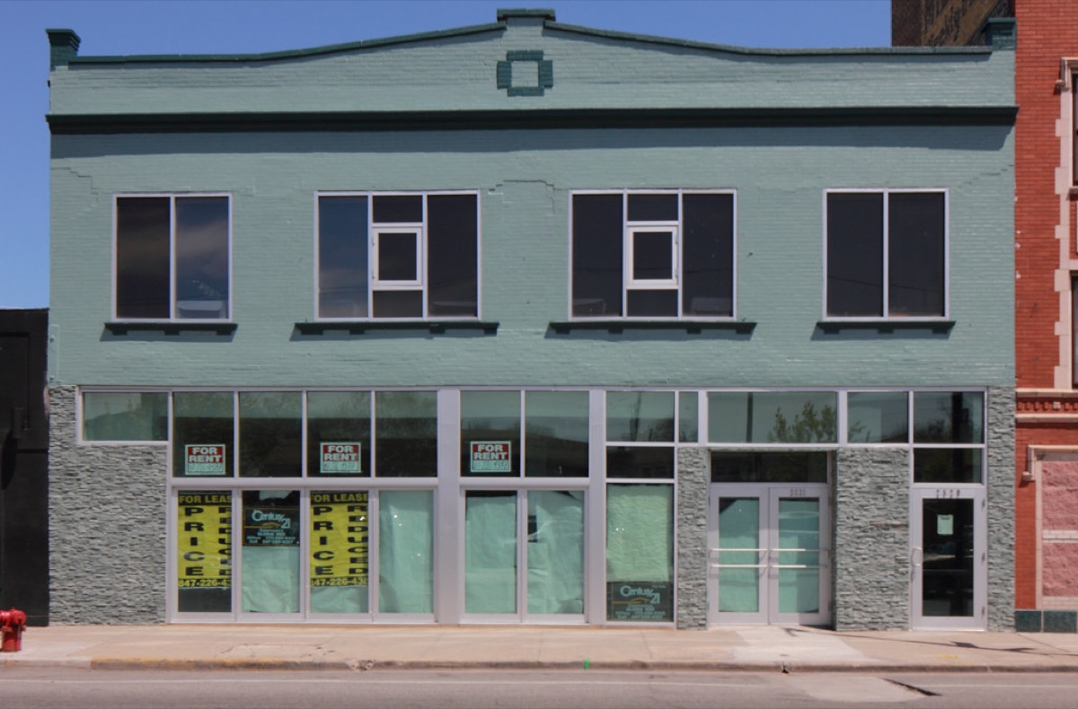 A vacant two storey commercial storefront with large ground floor windows, several red and white For Rent and yellow For Lease signs taped inside the glass, and a set of double entry doors.