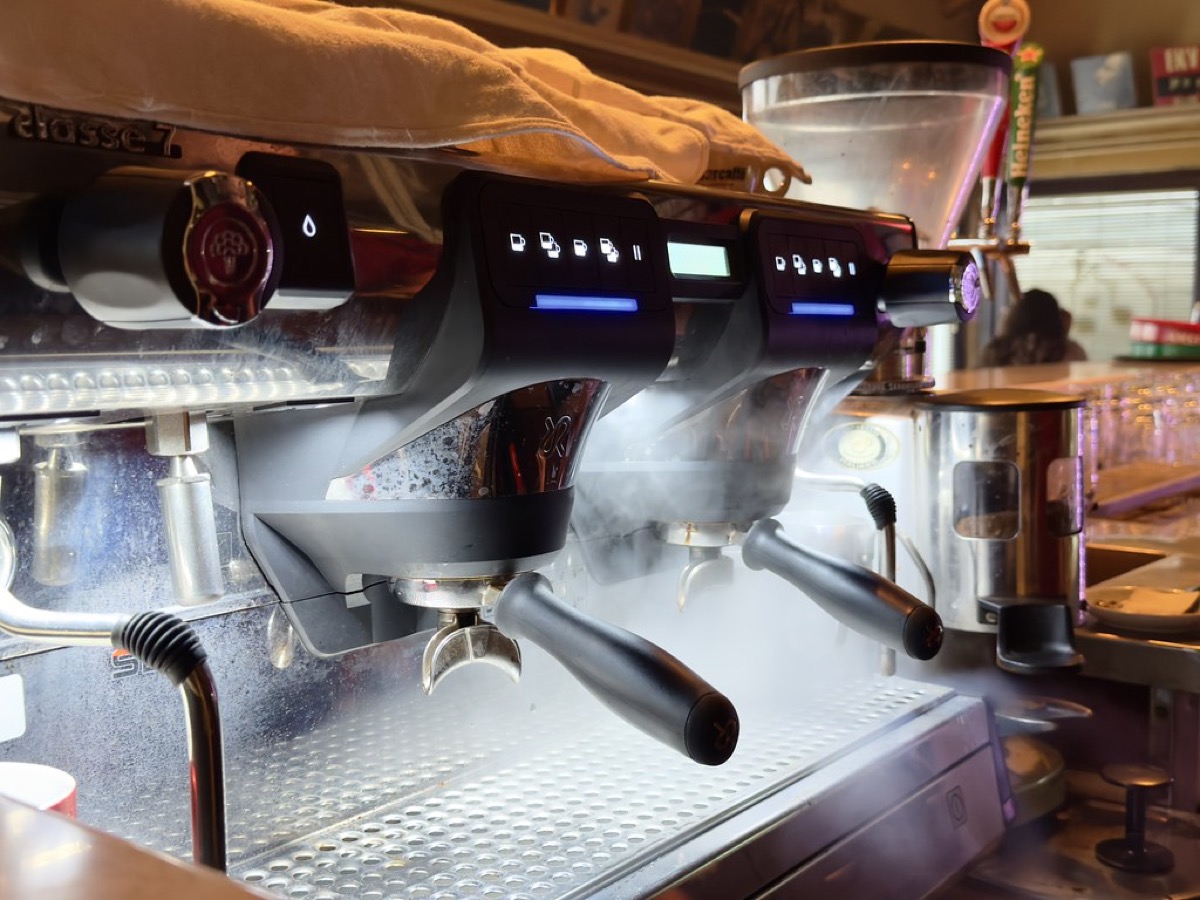 Close-up of a commercial two-group espresso machine with portafilters attached, steam rising from the group heads in a busy cafe.