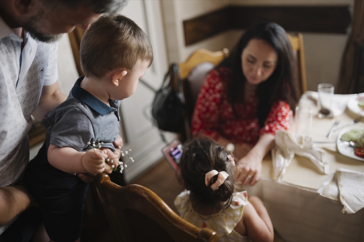 A father holding a toddler and a mother with a young daughter gathered around a set restaurant table with plates and glasses.