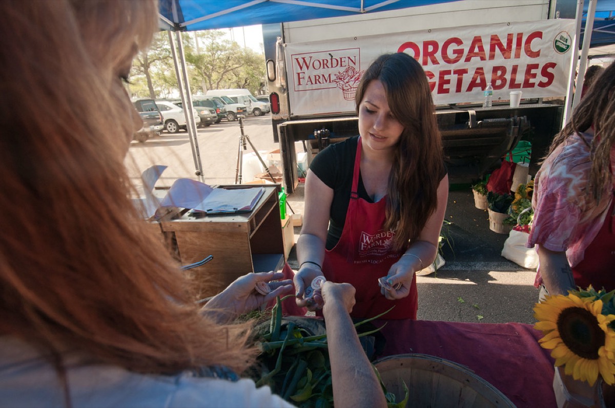 A customer hands cash to a vendor in a red apron at an outdoor farmers market vegetable stall, with sunflowers and produce baskets on the table.