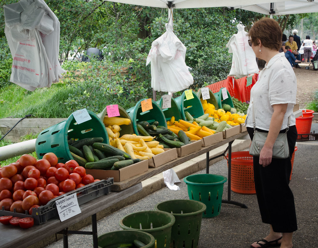 A shopper browses fresh tomatoes, cucumbers and squash at a farmers market stall under a white canopy