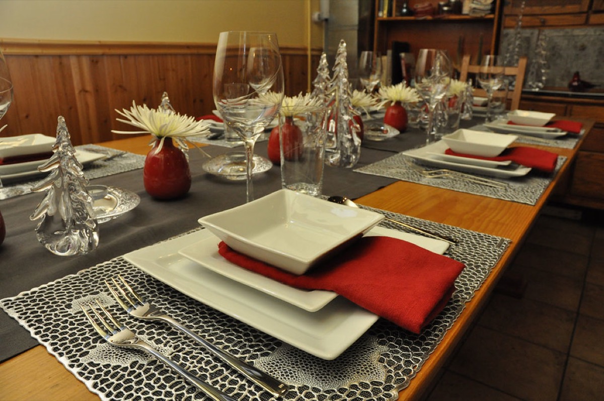 An elegant restaurant style table set for the holidays with white square plates, red napkins, wine glasses, glass Christmas trees and small red vases with white flowers