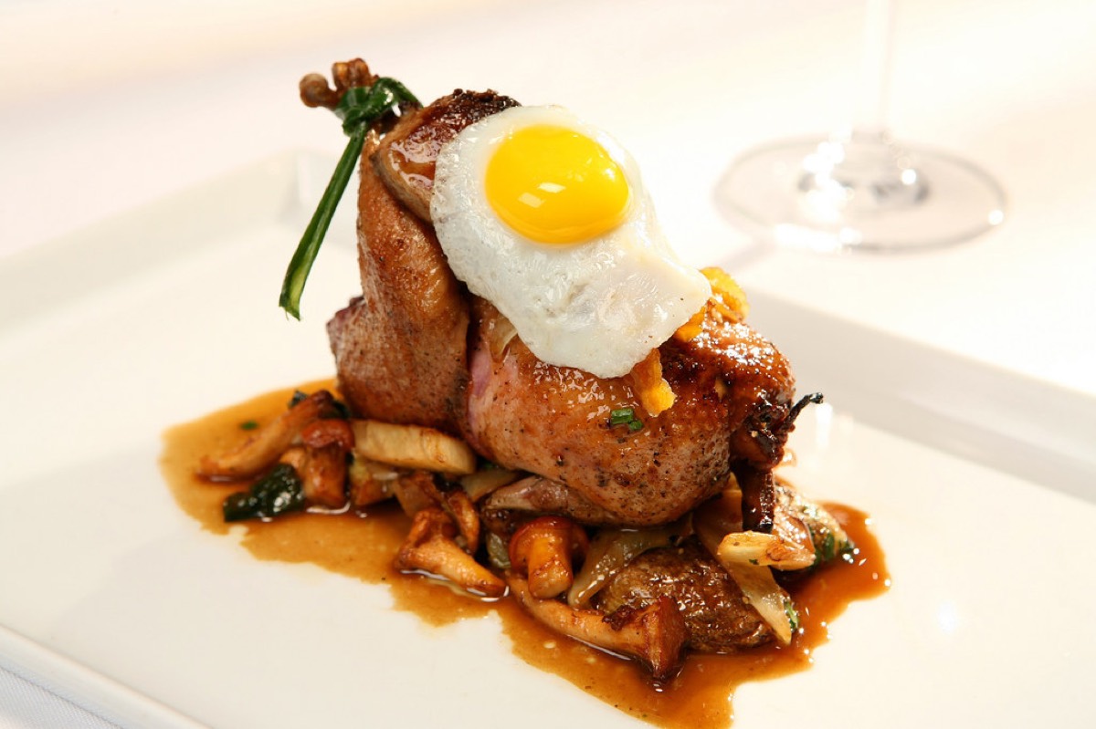 Seared quail topped with a fried quail egg, foie gras, and wild mushrooms on a white plate with a blurred wine glass behind