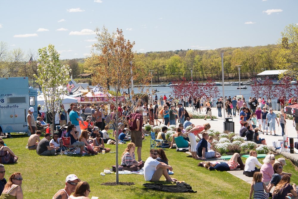 A large crowd relaxing on grass and walking along a riverside promenade at an outdoor food truck festival, with food trucks and tents in the background