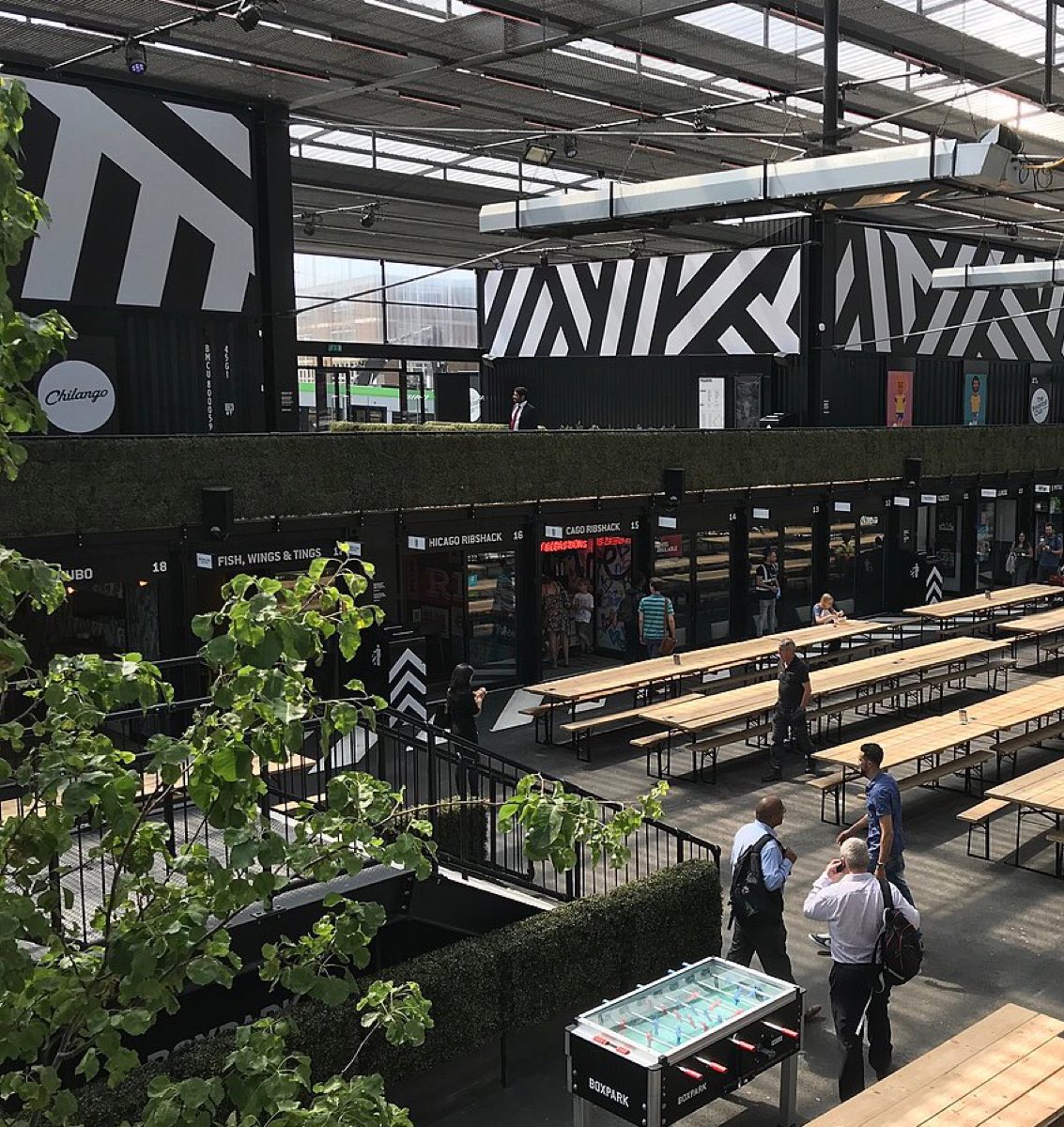 Daylit food hall with shipping-container food stalls, communal wooden picnic tables, and hanging greenery