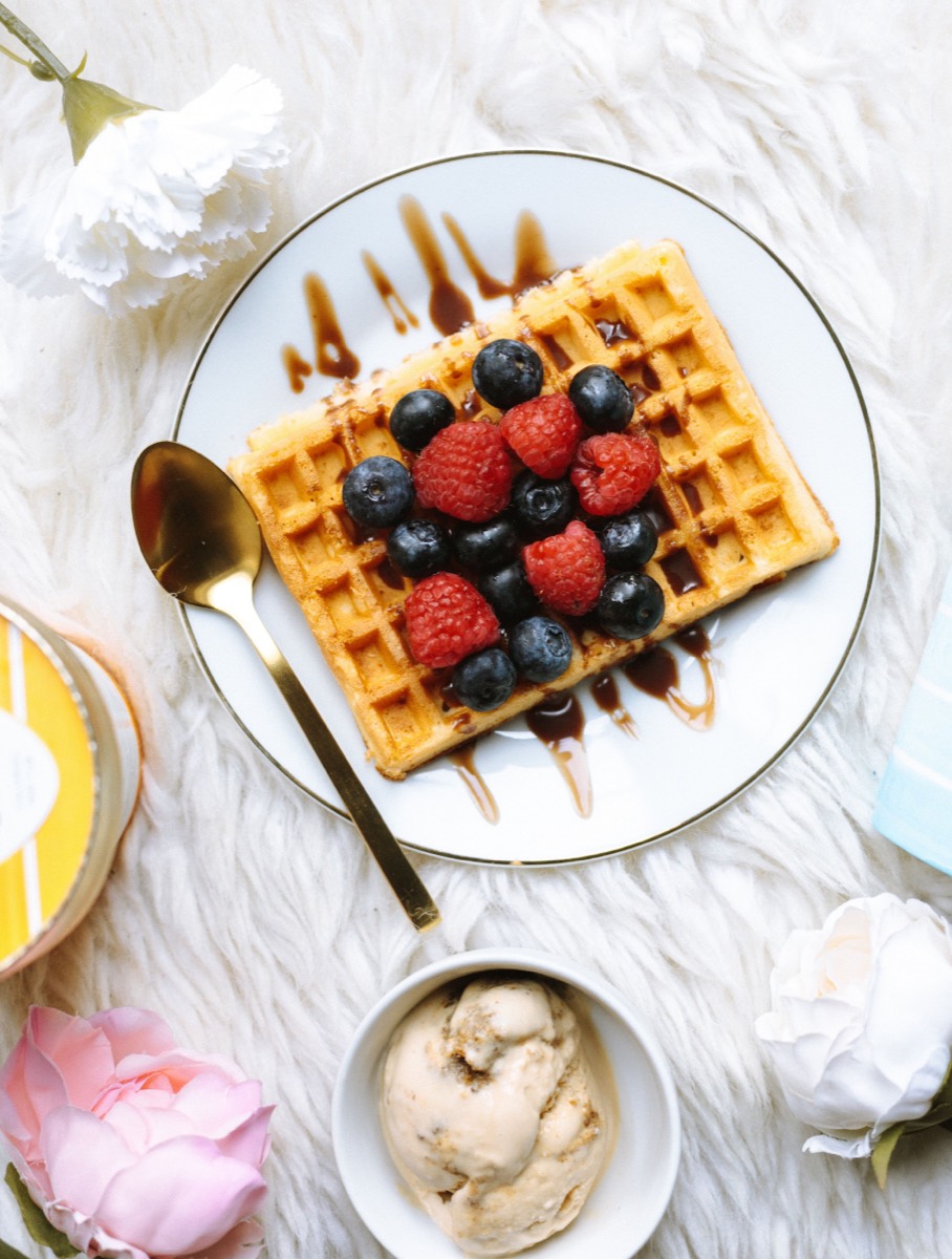 Overhead flat lay photo of a plated waffle topped with berries and chocolate syrup beside a bowl of ice cream