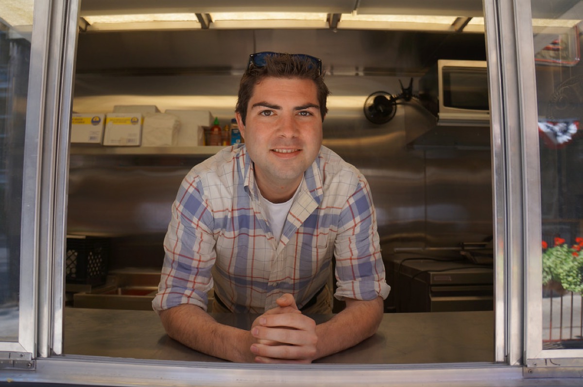 A food truck vendor leans on the stainless steel service window counter, smiling out at the camera with the truck's kitchen visible behind him.